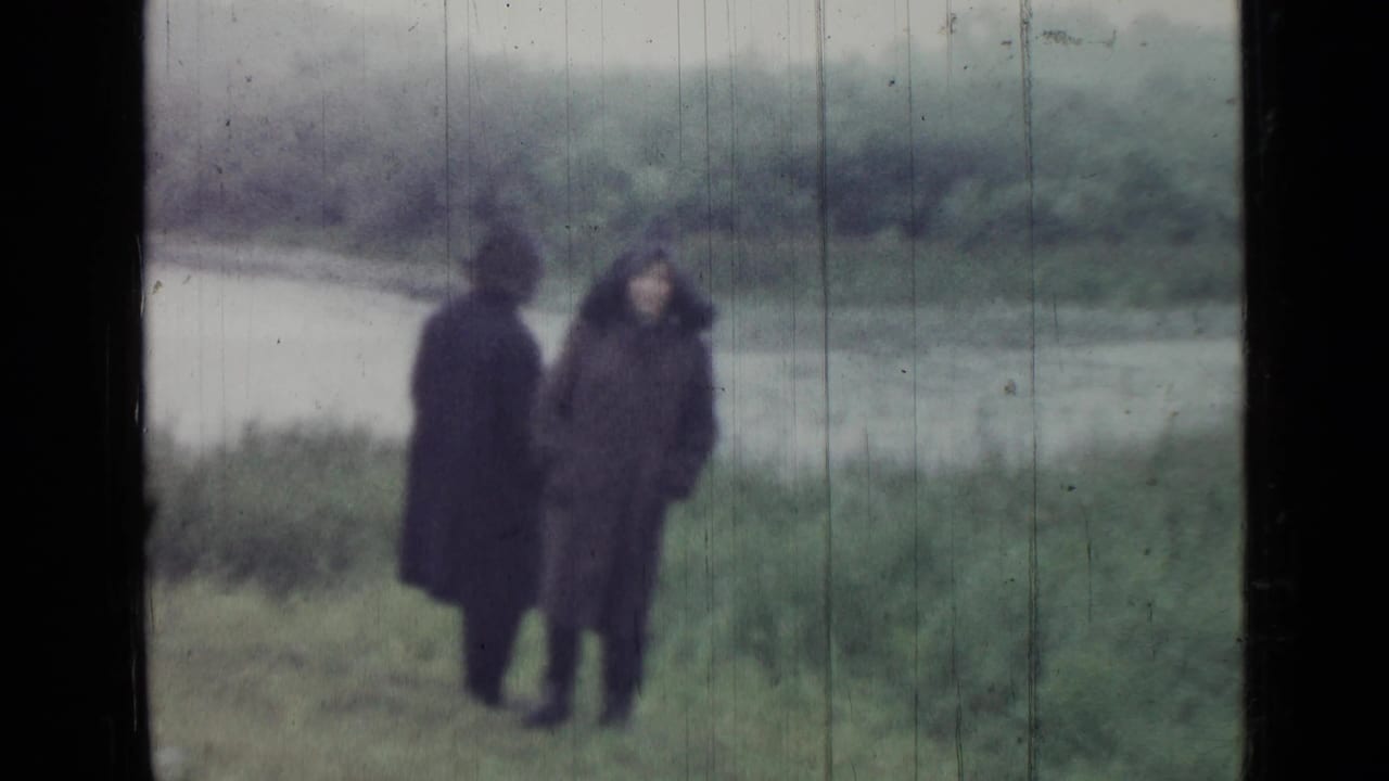 1967 Saint Petersburg frame with two coat-wearing adults standing near reeds and water under overcast light.