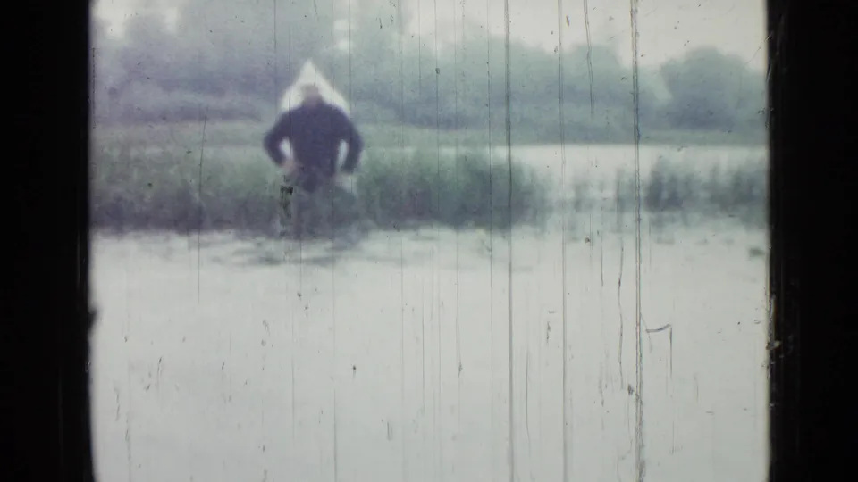 In this 1967 Saint Petersburg 8mm frame, a distant figure stands near reeds and water, partly obscured by a dark window frame and film scratches.