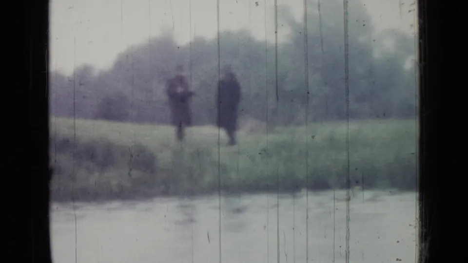 Two blurred figures appear to stand and chat on a grassy bank beside the lake in 1967 Saint Petersburg home-movie footage.