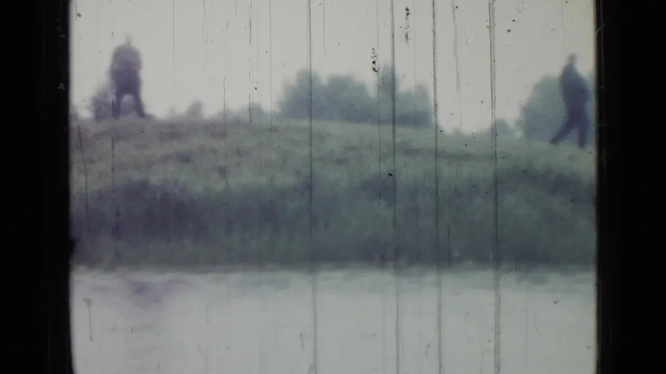 Distant people are visible on a raised grassy bank beside the lake, with soft focus and vertical scratches in the 1967 Saint Petersburg footage.
