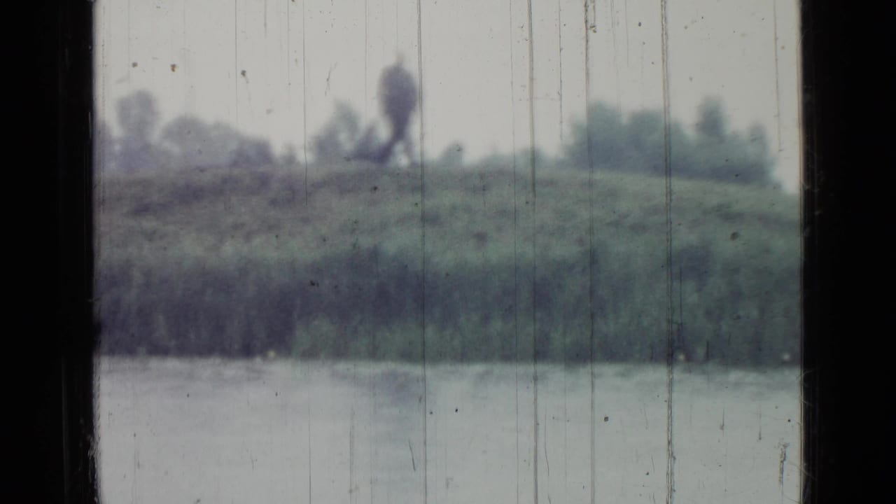 Distant figure walking along a grassy bank across a pond, seen through scratched rain-streaked glass.