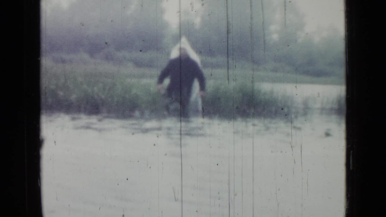 A distant figure in a light hat and dark coat wades through marsh water in the 1967 Saint Petersburg home movie.