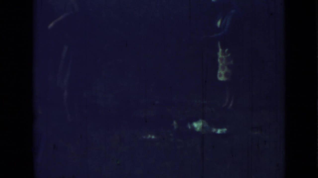 Blue-tinted 1967 home-movie frame showing indistinct silhouetted figures, reflective highlights, dark borders, and visible film wear.