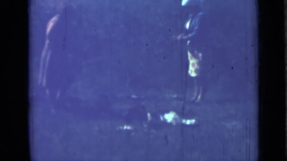 1967 8mm frame with a dim standing figure, possible patterned clothing, blue color cast, reflective spots, and heavy film scratches.
