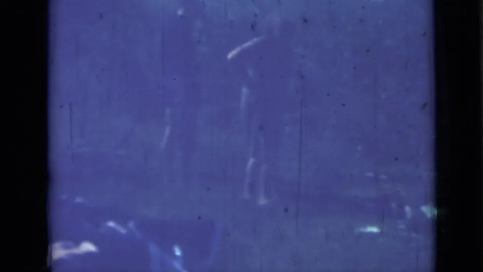 Dim 1967 8mm frame showing a faint standing human silhouette, blue exposure, scratches, dark border, and blurred background forms.