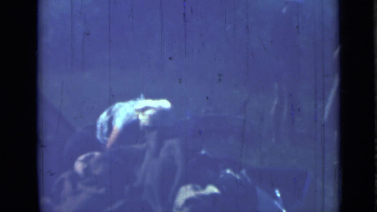 Blue-tinted 8mm frame with women and camp equipment in a dim outdoor setting.