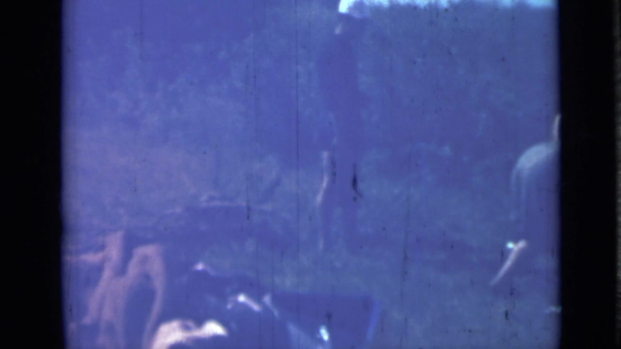 Dim blue frame with figures near a tent or camp structure and outdoor equipment.
