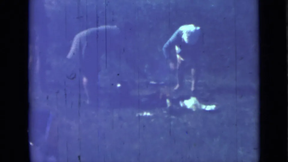 A woman bends beside camp cooking items in the 1967 Saint Petersburg-area rural footage, with tents and figures faintly visible.