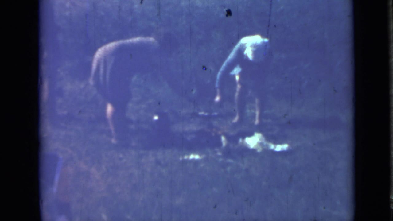 Two women bend over a cooking area in a blue-tinted wooded campsite frame.