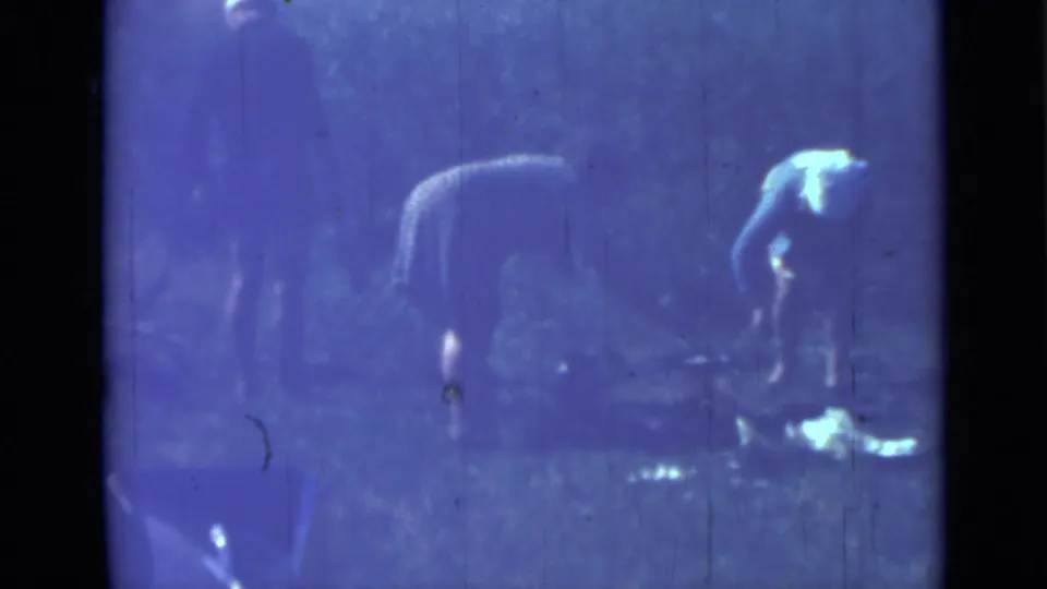 A blue-shifted 1967 8mm frame shows one woman upright and another bending near a low cooking area in a wooded Saint Petersburg campsite.