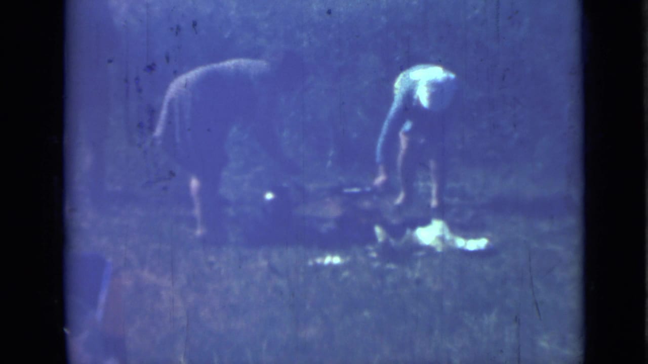 Two women bend near the ground in a blue-tinted outdoor camp scene.