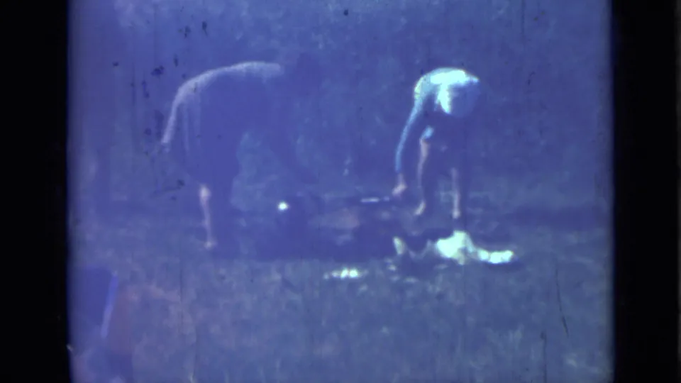 1967 home-movie frame from Saint Petersburg area showing women bent over outdoor meal preparation in a grassy camp setting.