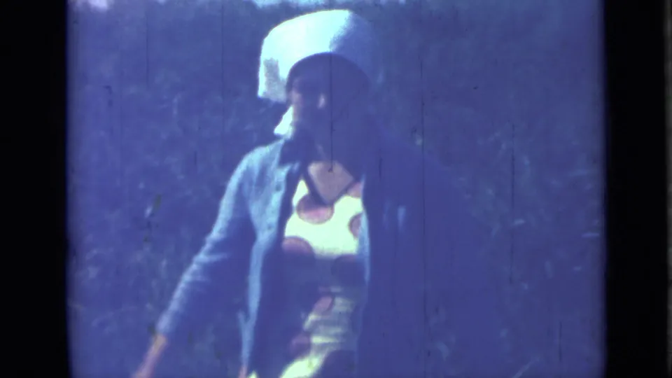 1967 Saint Petersburg home-movie frame showing a woman in a white headscarf, patterned dress, and cardigan at an outdoor camp.