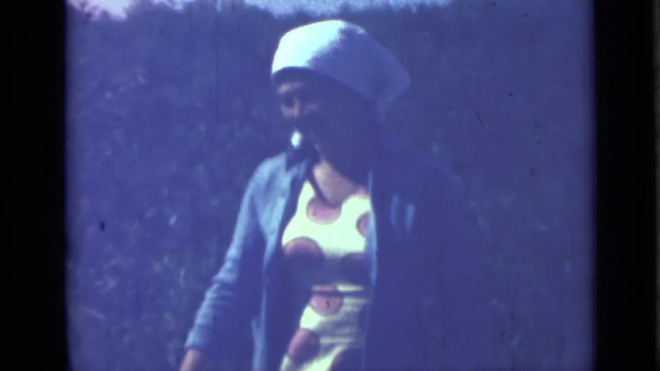 1967 Saint Petersburg 8mm frame of a woman in a white headscarf and cardigan turning in an outdoor camp setting.