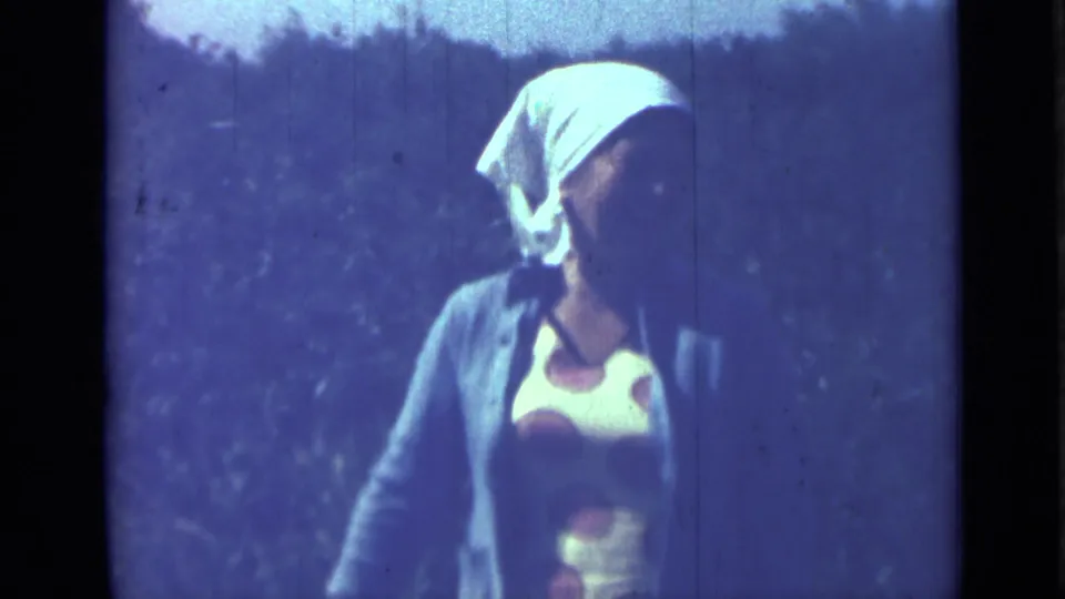A woman in a white head covering and polka dot dress turns slightly during the handheld 8mm shot.