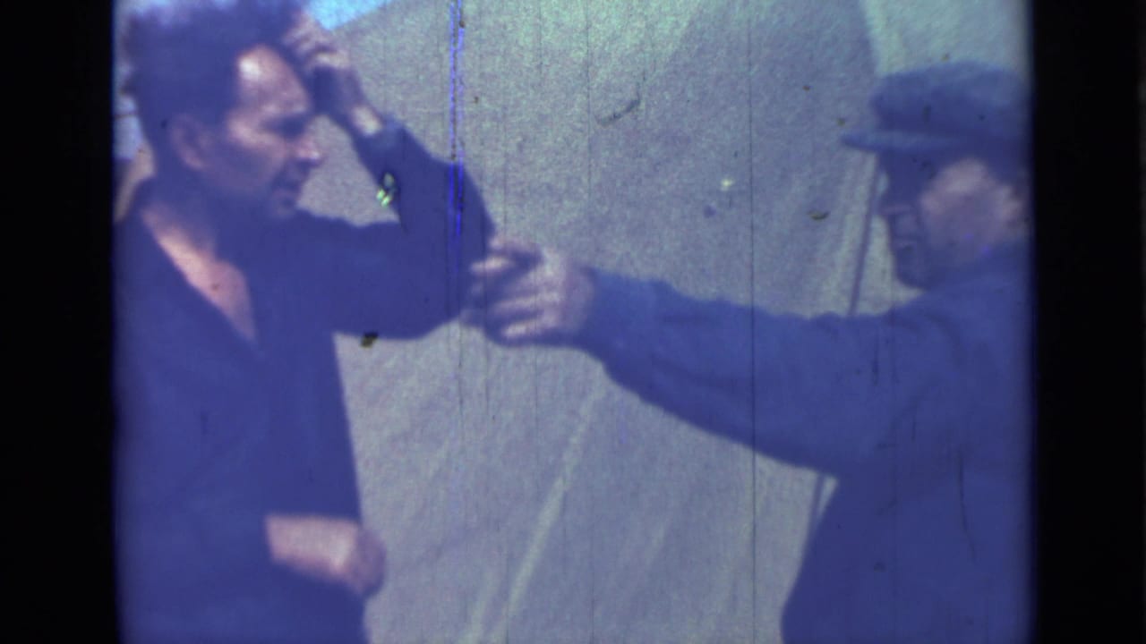 Two adult men outdoors, one in a dark shirt and one in a cap, gesturing close together.