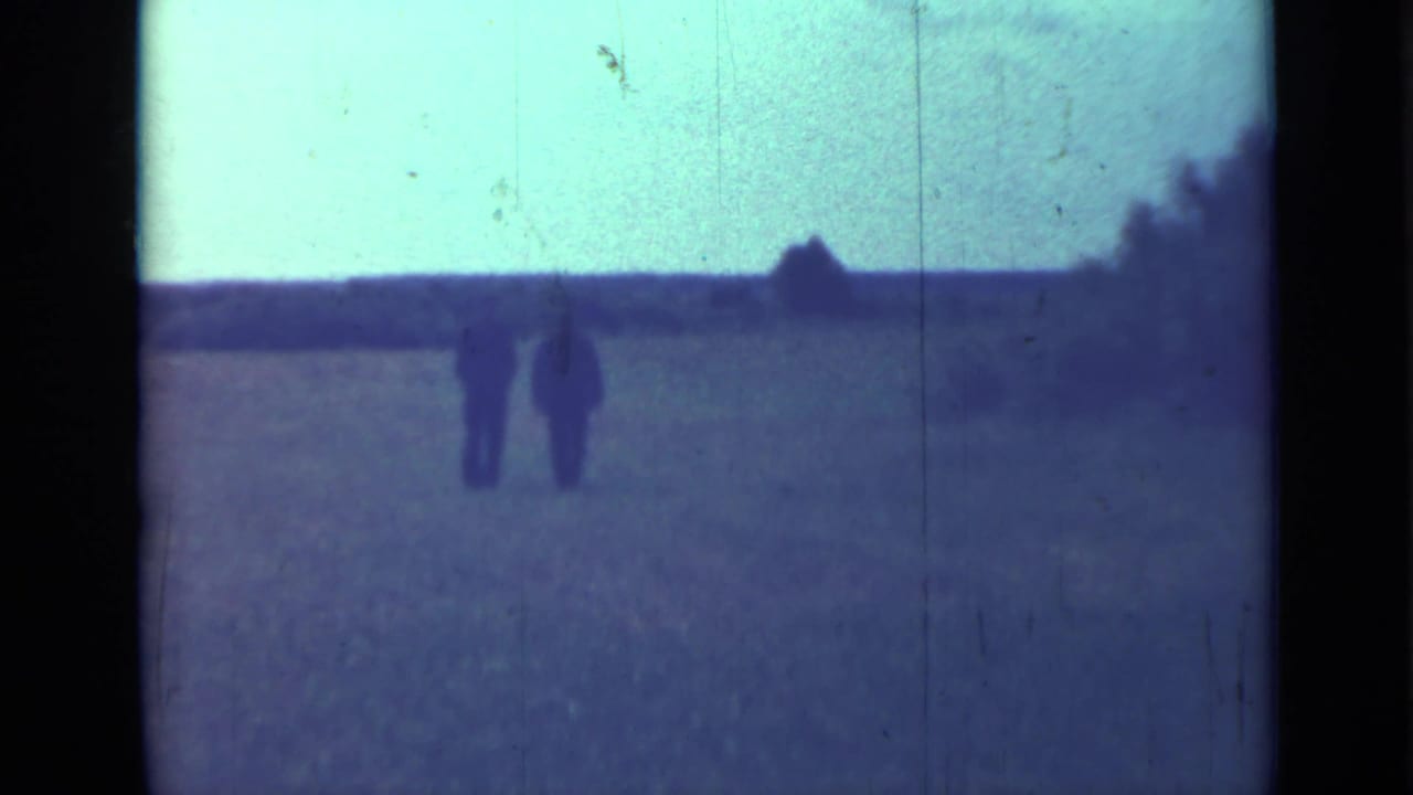 Two distant men walk across an open field beneath a pale sky.