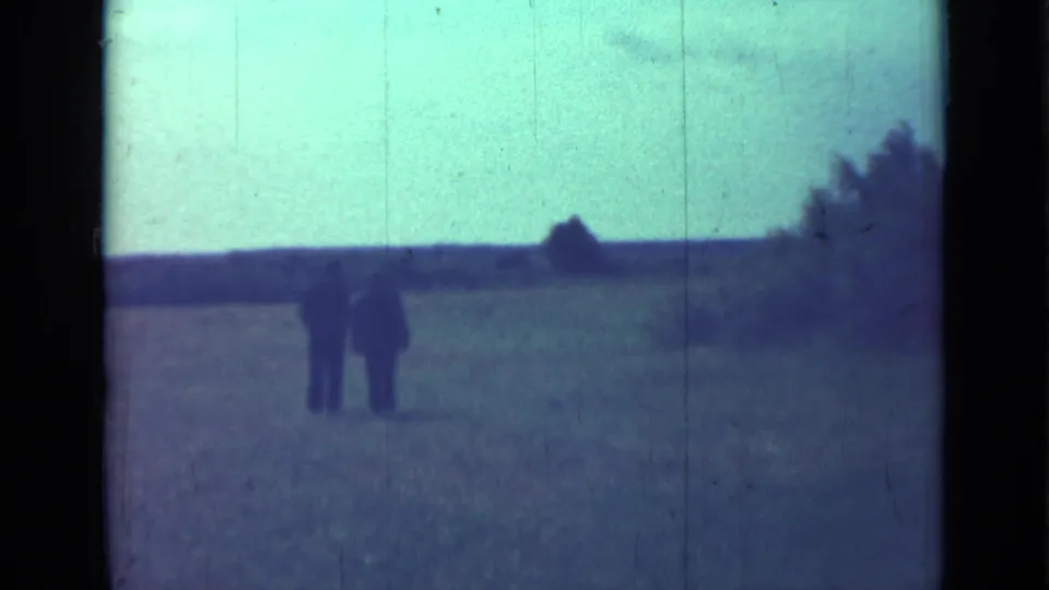 A soft 8mm frame shows two men walking away through an open field near Saint Petersburg, with a dark horizon line beyond them.