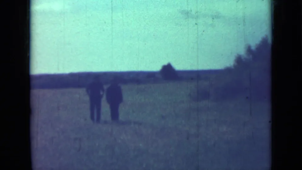 Final 1967 home-movie frame shows two distant men walking in a field, with trees and a small horizon structure or mound behind them.
