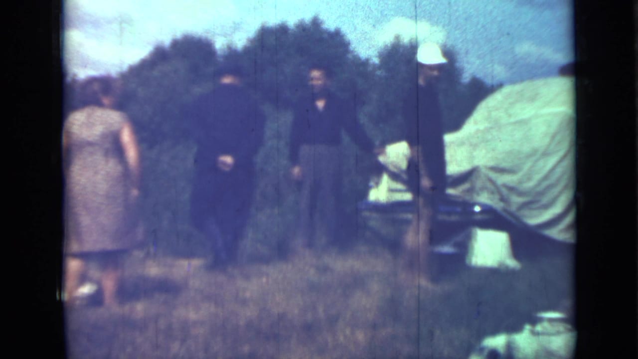 Adults stand in tall grass near a covered vehicle, with trees and sky behind them.