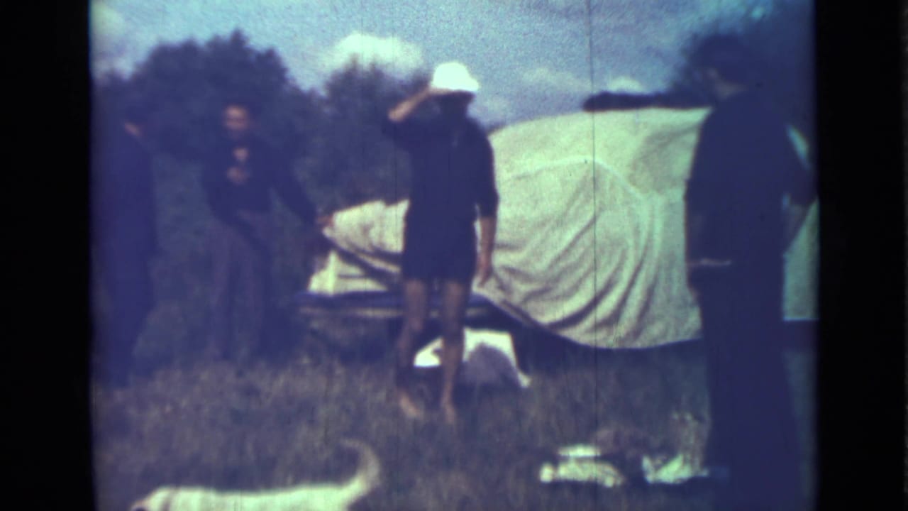 A person in a white hat stands by a covered vehicle while others remain nearby in grass.