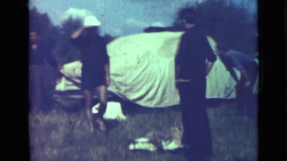 1967 campsite frame with adults near a covered vehicle and a small dog crossing or standing near the bottom of the shot.