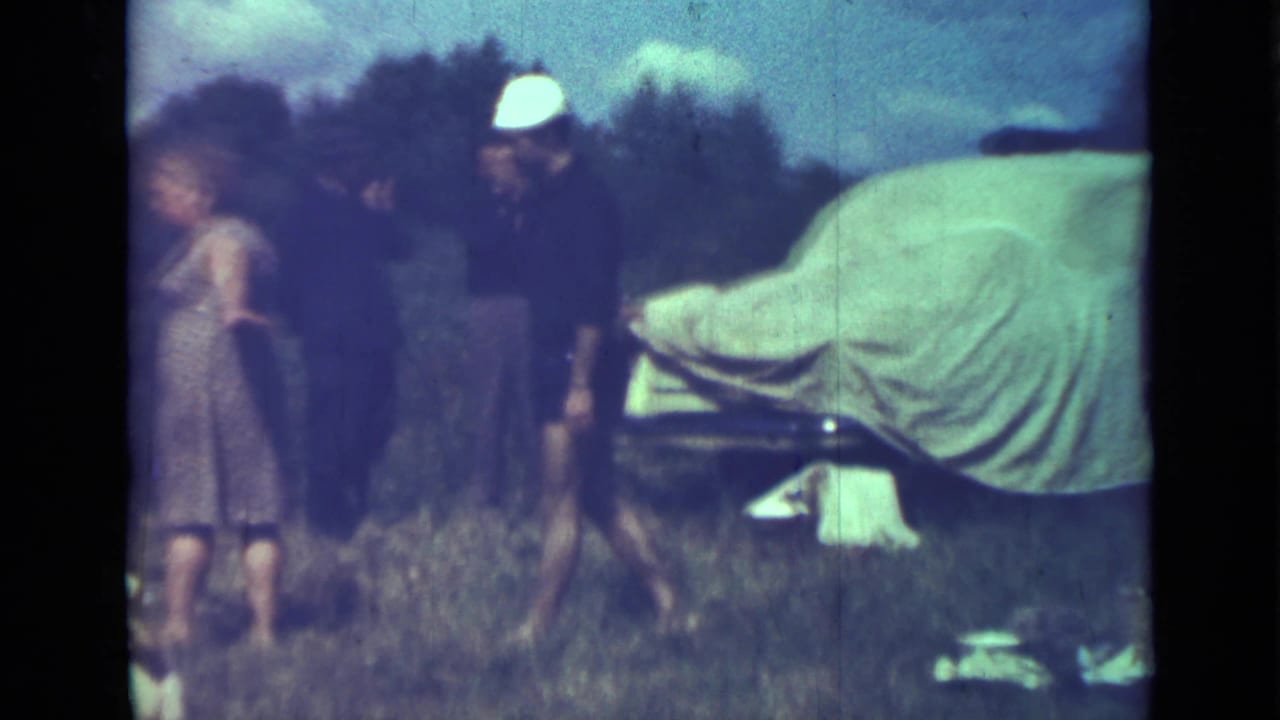 Adults stand in grass near a canvas-covered car, with trees in the background.