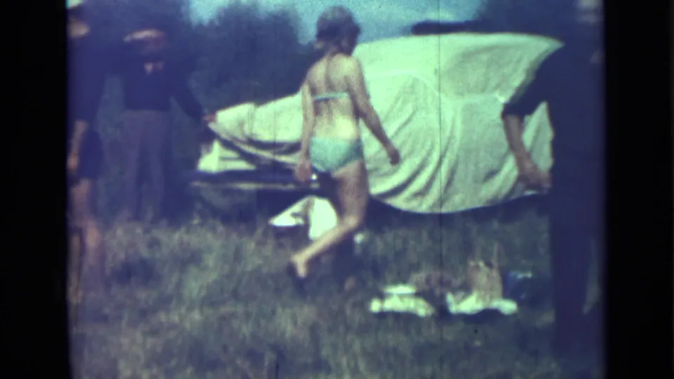 A woman in a swimsuit crosses the frame during a 1967 Saint Petersburg home-movie scene beside a canvas-covered car.