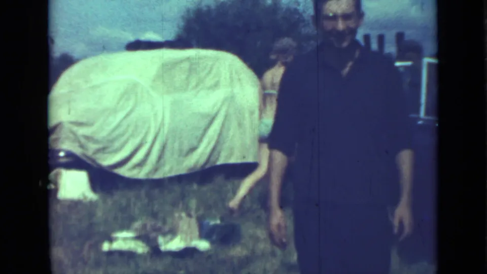 A man faces the camera in a soft 1967 8mm frame, with a canvas-covered car and other adults behind him.