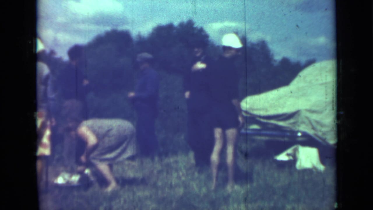 Adults gather in a grassy field near a covered car while one person crouches by the ground.