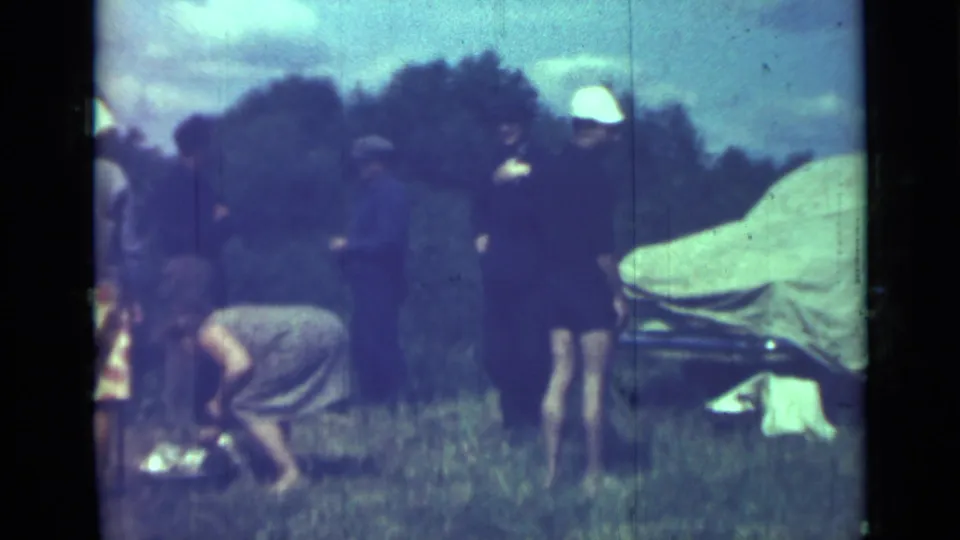 1967 home-movie frame near Saint Petersburg showing adults in a grassy camping area with a covered car and someone crouching near food or gear.