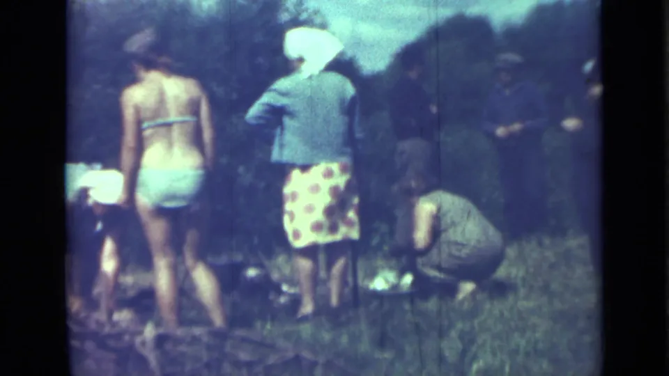 Saint Petersburg 1967 home-movie frame showing adults in summer clothing around a grassy campsite, including a person bending near the ground.