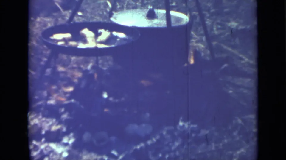 1967 Saint Petersburg home-movie frame showing cast iron cookware over an open outdoor fire, tinted blue-purple by aged 8mm film.