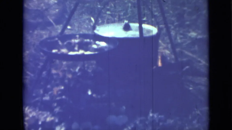 Blue-purple 8mm frame from 1967 showing a pan, larger pot, and open flame in an informal Saint Petersburg outdoor cooking scene.
