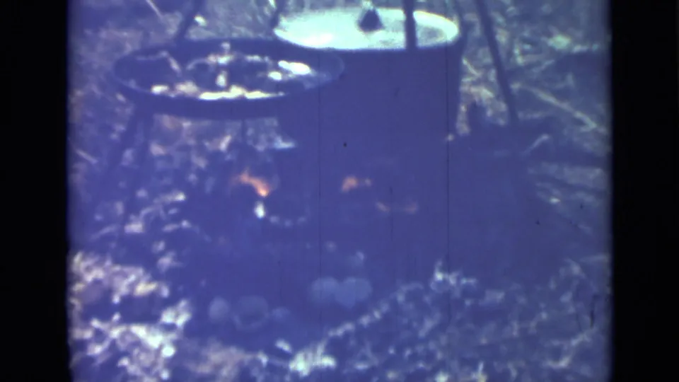Aged 8mm frame of the Saint Petersburg outdoor cooking setup, with dark cookware, fire glow, rough ground, and visible film grain.