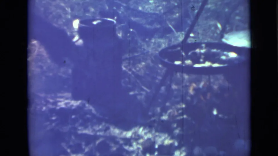 A partly visible hand or arm tends the tripod cookware in this 1967 Saint Petersburg home-movie frame.