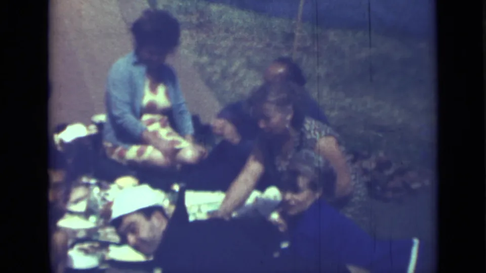 Adults sit around picnic blankets and food in a 1967 Saint Petersburg home-movie frame, with handheld 8mm softness and color shift.