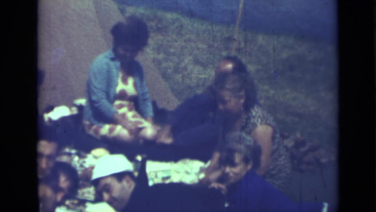 Adults seated on the ground around picnic food in a soft 8mm home-movie frame.