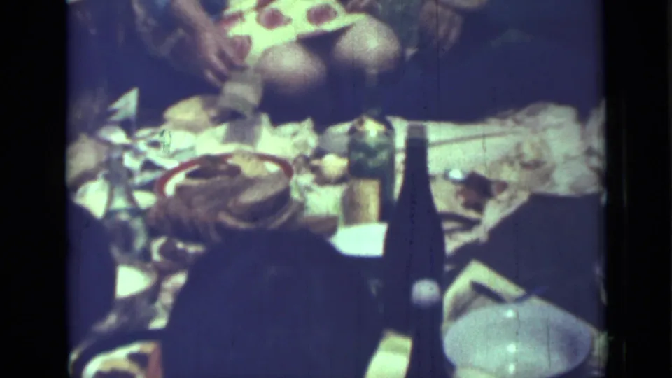 Saint Petersburg 1967 home-movie frame of a picnic-style meal with dishes, bottles, food, and seated people nearby.