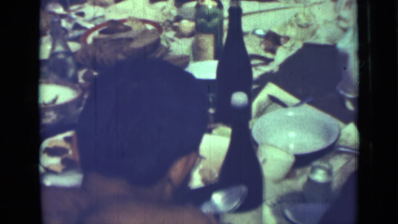Shirtless man seen from behind at a crowded dining table with bottles, bowls, plates, and food.