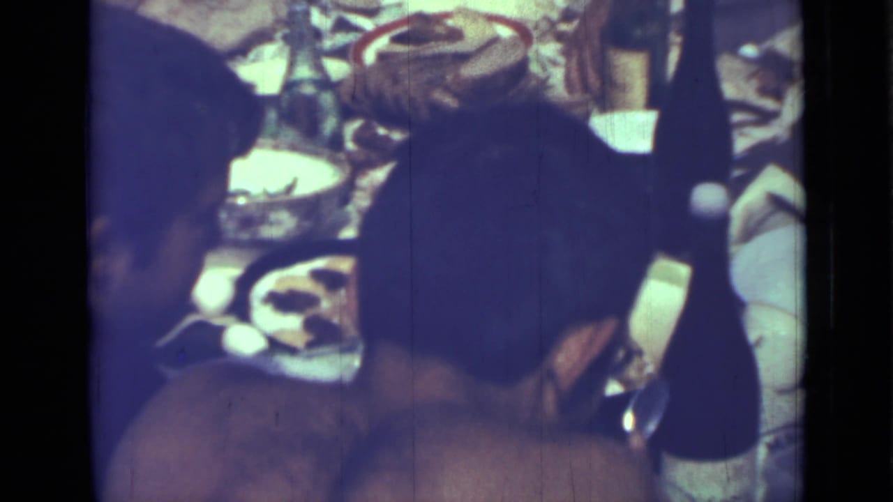 Two adults sit near a crowded table with dishes, bottles, and food, one seen mostly from behind.