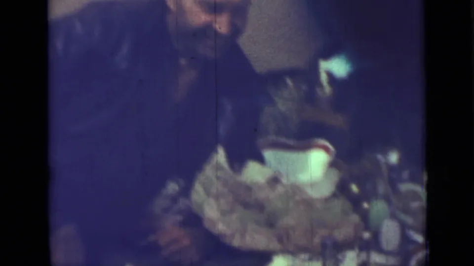 1967 Saint Petersburg home-movie frame showing an adult in a dark jacket leaning over a dining table with dishes and a bottle.