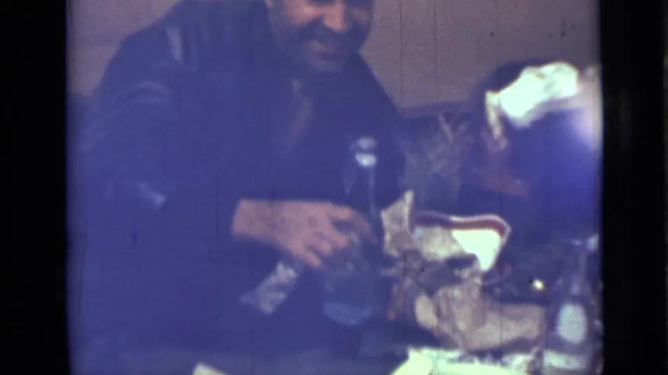1967 Saint Petersburg home-movie frame showing a seated man in a dark jacket at a dining table with a bottle, dishes, and a pale hat or cloth nearby.