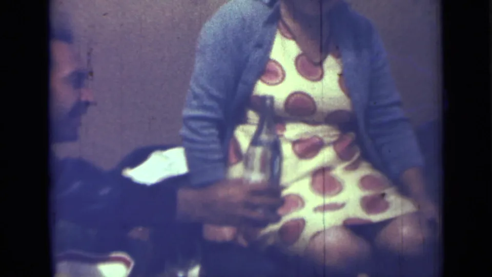 1967 Saint Petersburg 8mm frame showing a seated woman in a patterned dress beside a hand holding a bottle.