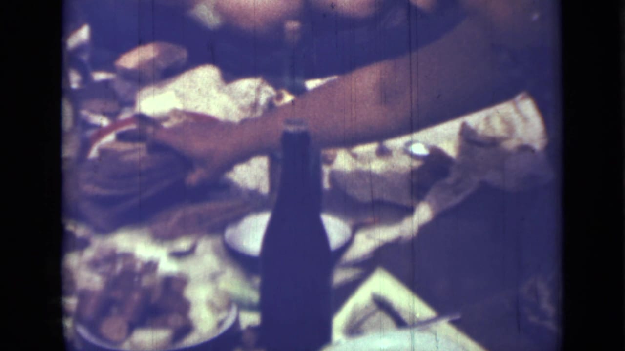 Crowded picnic table with bread, dishes, a dark bottle, and an adult arm reaching across.