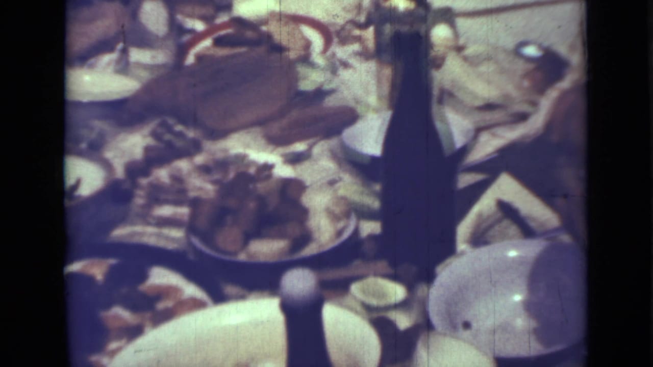 Picnic table with food, plates, a woven basket, dark bottle, and pale serving dish.