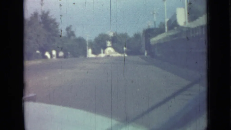 1967 Saint Petersburg home-movie frame looking from a vehicle toward a wide road, sidewalk, trees, and a distant white monument.