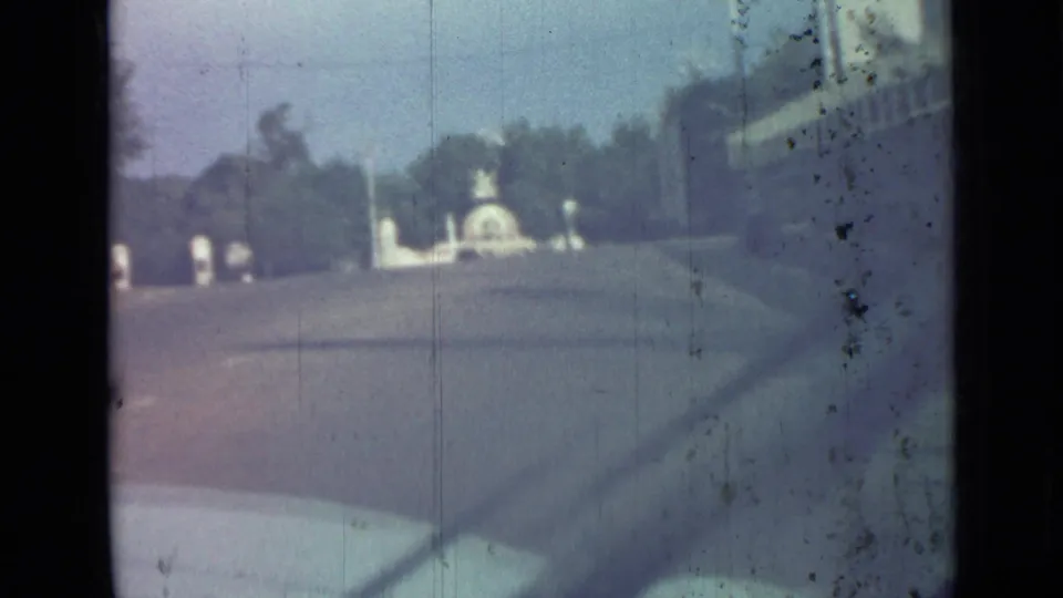 1967 Saint Petersburg home-movie frame showing a broad roadway from a moving car, with trees and a pale monument ahead.