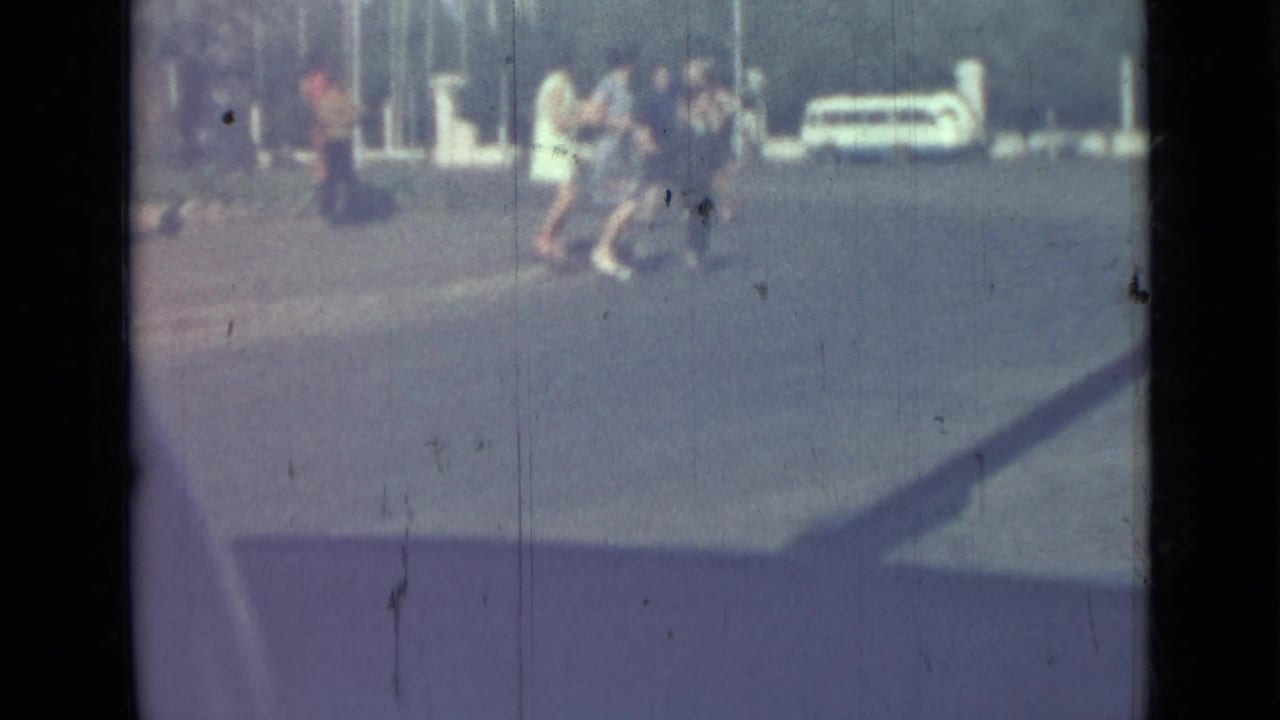 Distant pedestrians cross a sunlit paved lot near parked cars and white posts.