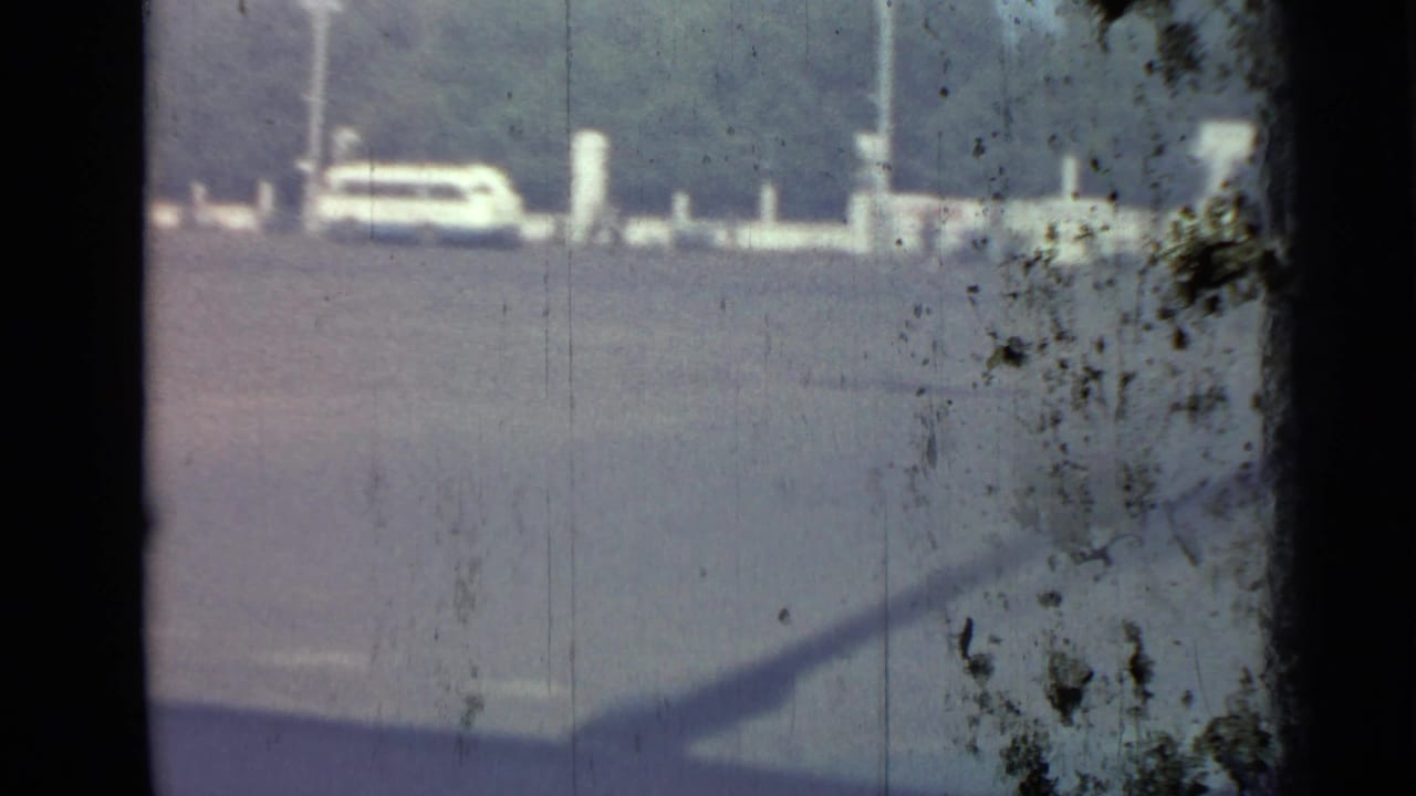 A pale parked car sits beside white posts at the edge of a broad paved area.